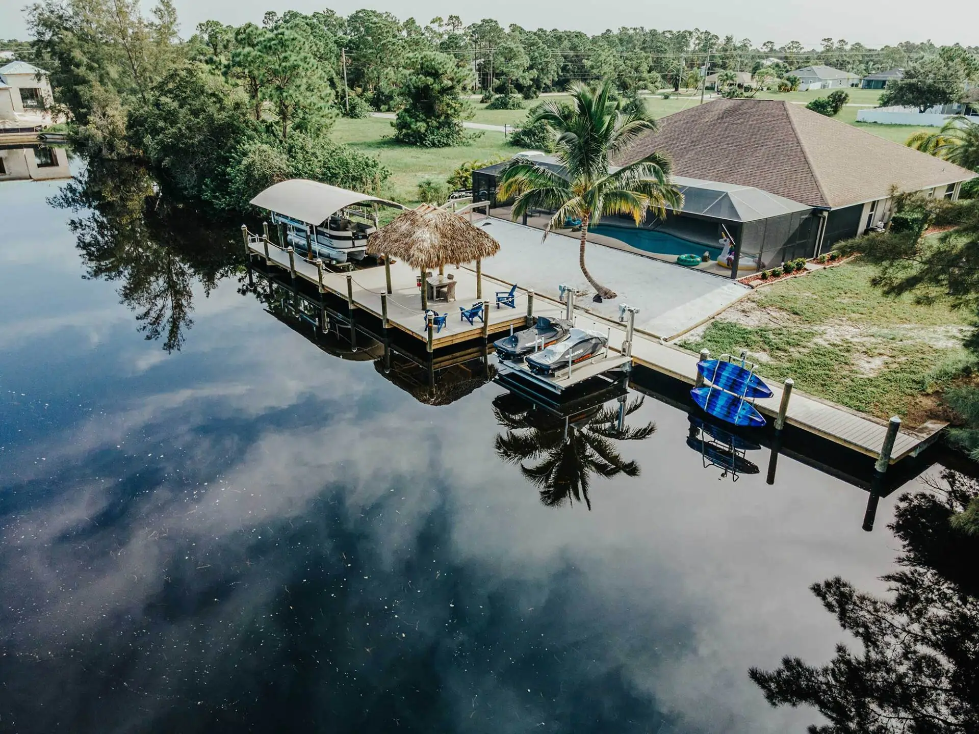 Aerial view of a residential dock with a tiki hut, covered boat lift, and paddleboards near a peaceful waterfront.