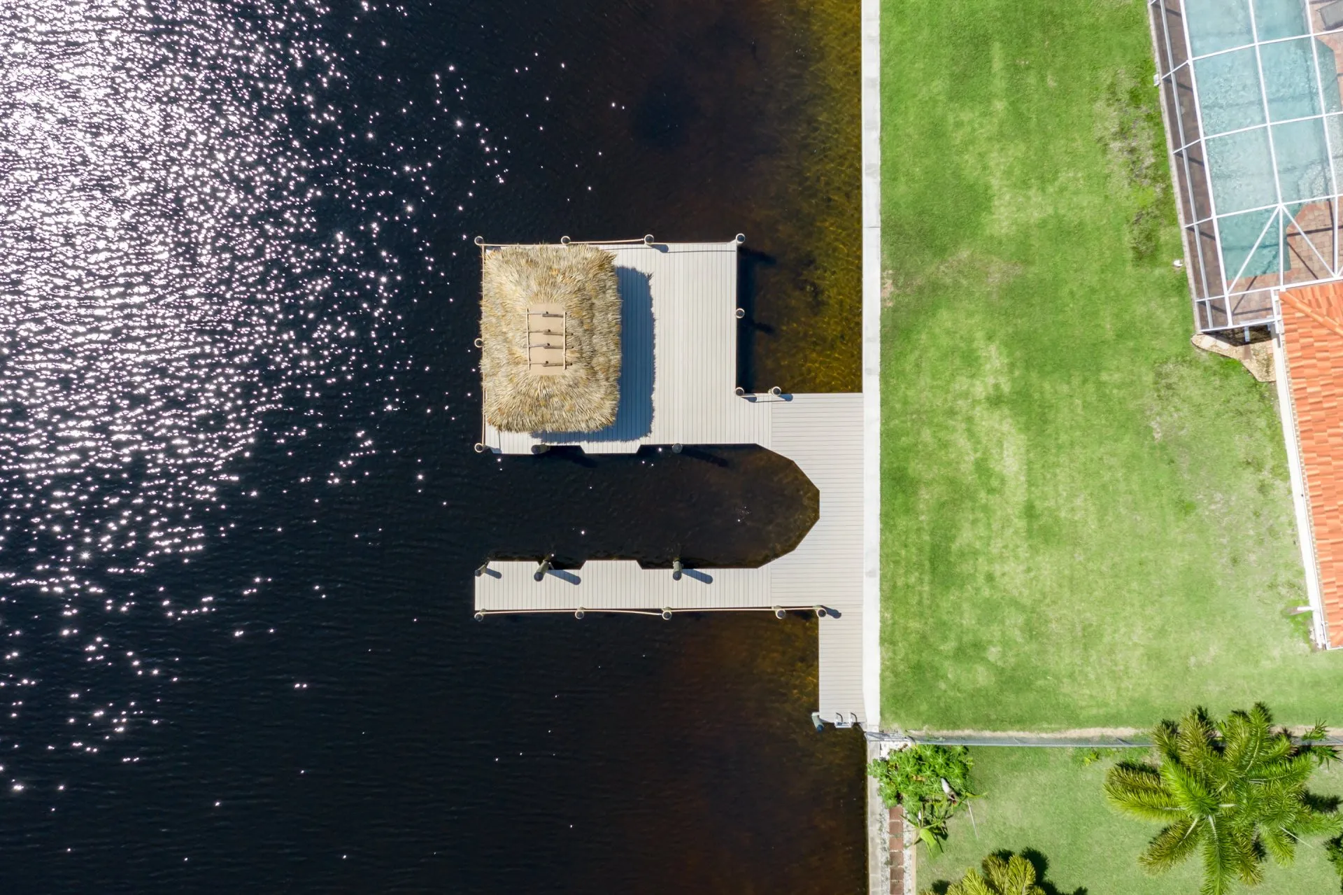 Aerial view of a dock with a tiki hut over a shimmering canal next to a grassy yard.