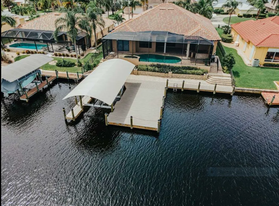 Aerial view of a canal dock with a curved canopy, composite decking, and a waterfront home with a screened pool.