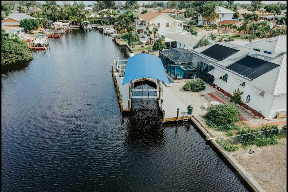 Aerial view of a canal dock with a blue curved canopy, boat lift, and a waterfront home with a screened pool.