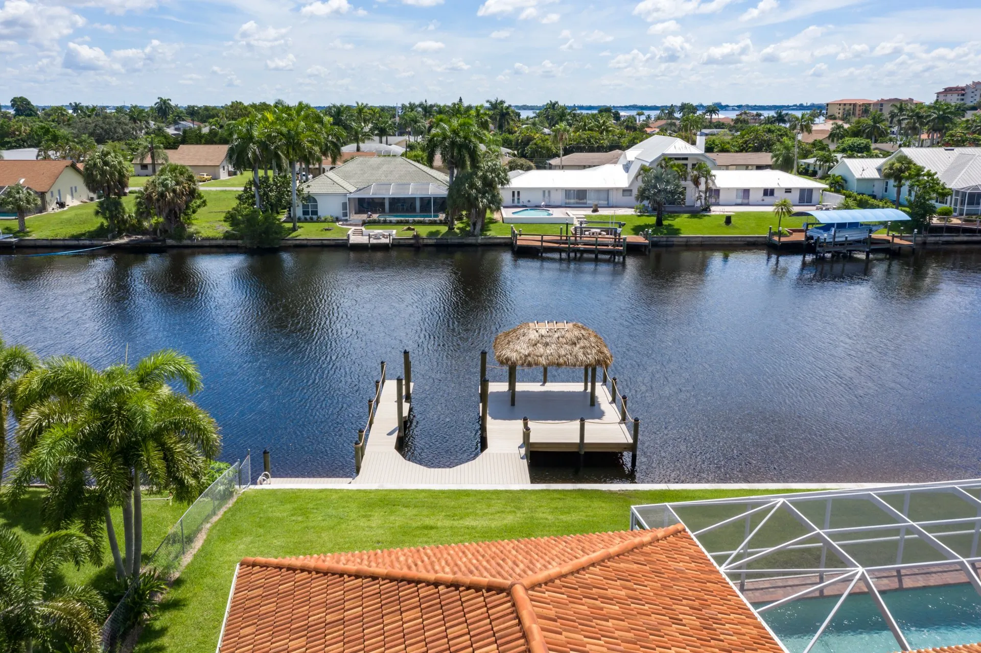 Aerial view of a tiki hut dock extending into a canal surrounded by waterfront homes and lush landscaping.