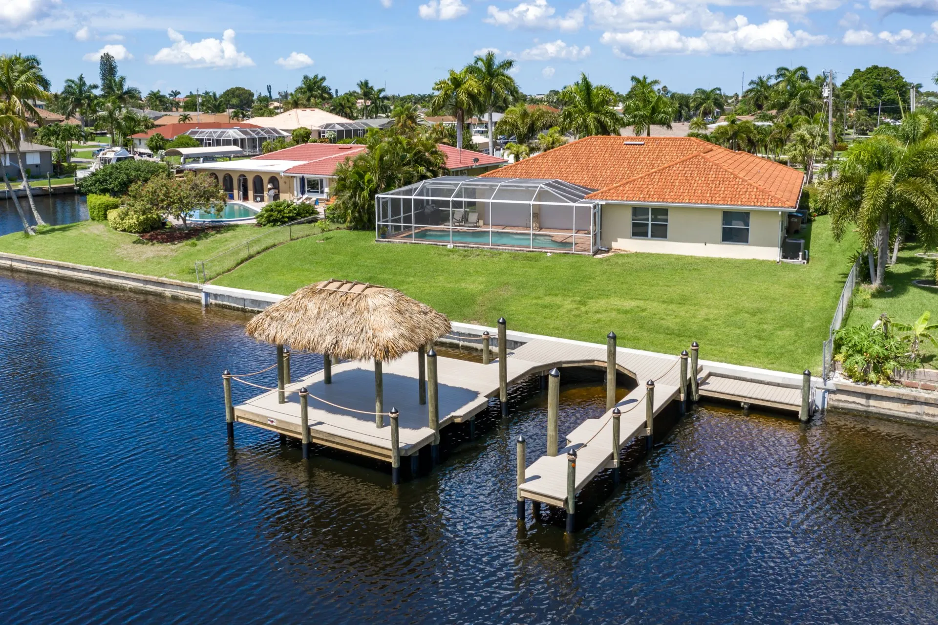 Elevated view of a backyard with a dock, tiki hut, and a screened pool area in a suburban waterfront neighborhood.