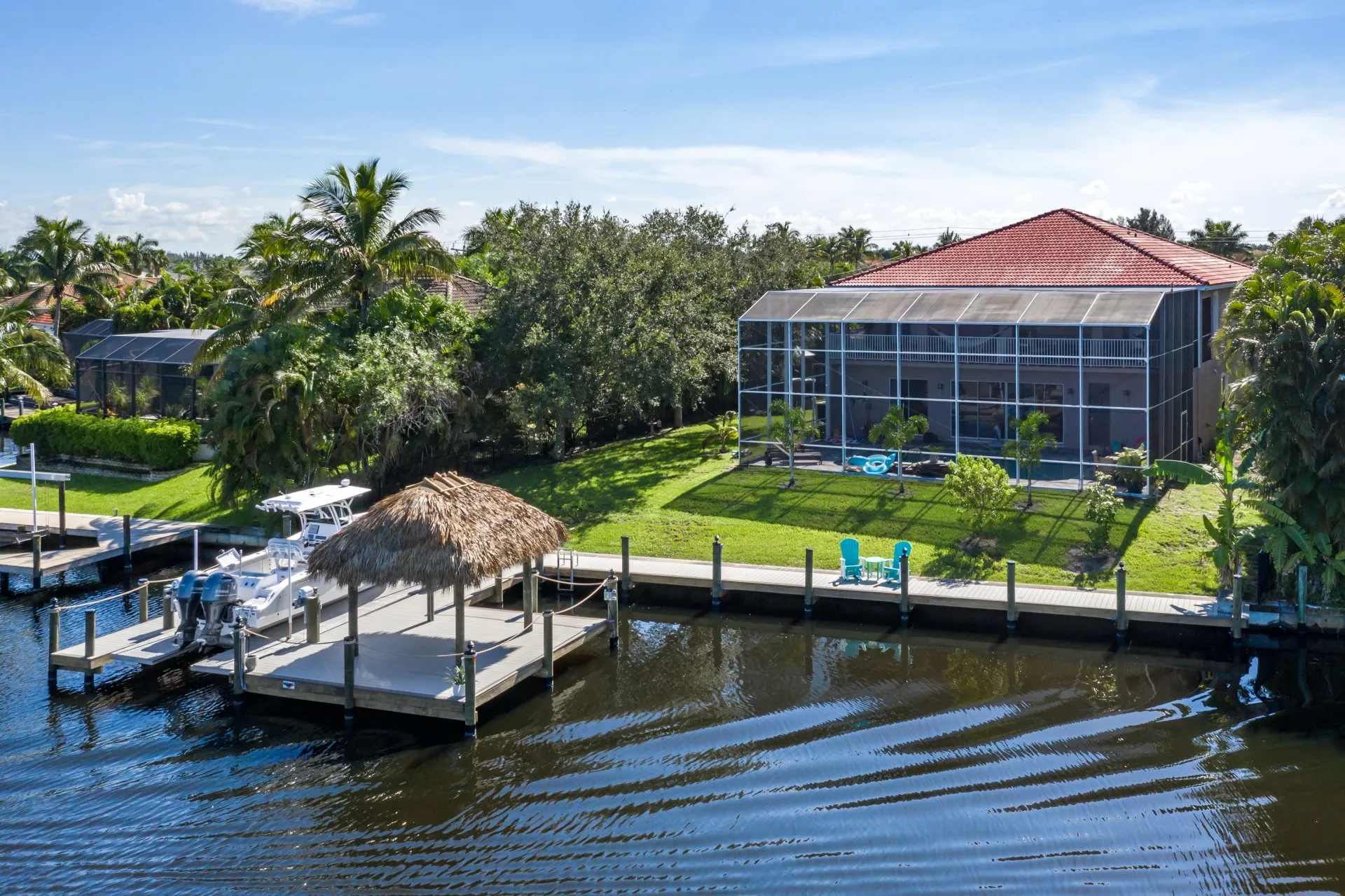 A scenic backyard featuring a screened lanai, lush greenery, and a waterfront dock with a tiki hut.