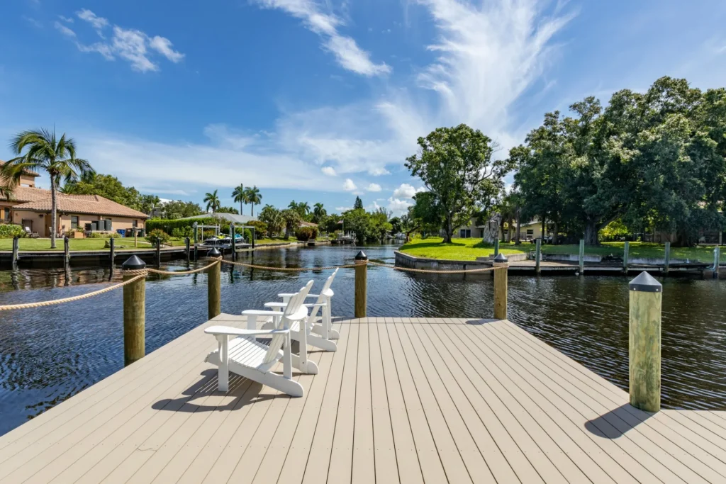 A waterfront deck with white chairs overlooking a peaceful canal surrounded by greenery and houses.
