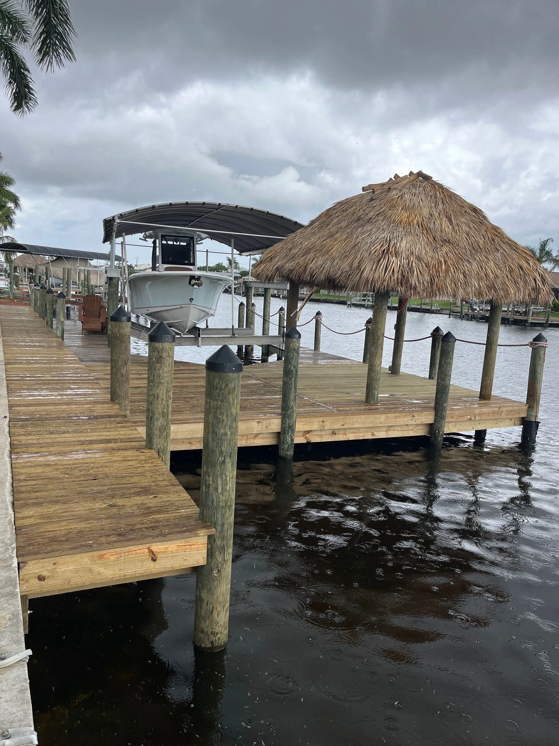 Canal dock with a thatched tiki hut, boat lift under a curved canopy, and wooden decking on a rainy day.