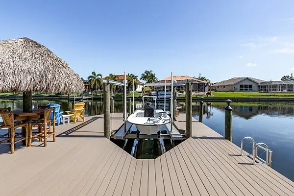 A canal-side dock featuring a boat lift with a small boat, a tiki hut, and colorful seating in a residential setting.