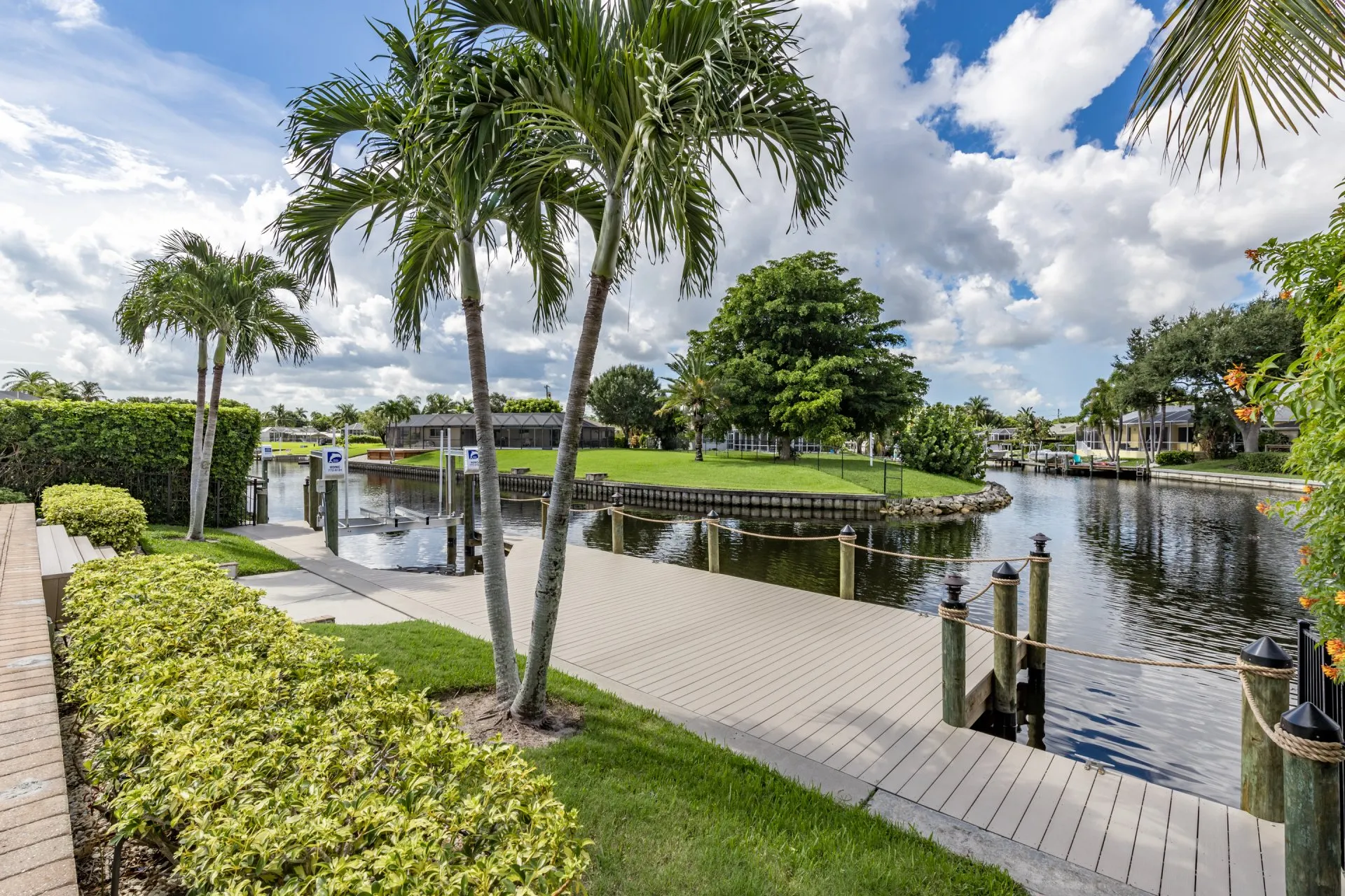 A canal-side dock with wooden posts, rope railing, and surrounding palm trees and greenery under a partly cloudy sky.