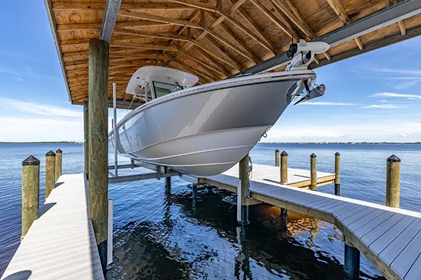 A boat lifted under a wooden cover at a waterfront dock, surrounded by calm waters and a clear sky.