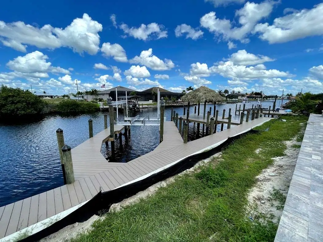 Curved wooden dock leading to a tiki hut on calm water under a vibrant blue sky with fluffy clouds.