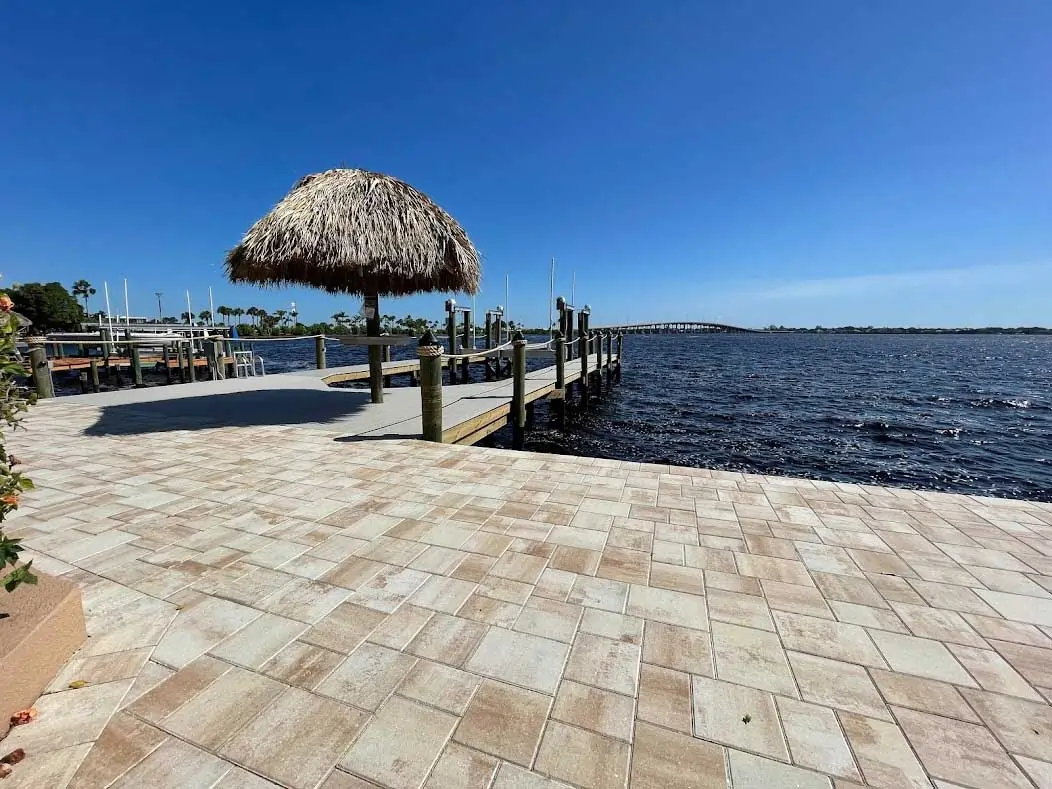 Tiki hut on a dock overlooking a wide waterfront with a paver walkway and a distant bridge under clear skies.