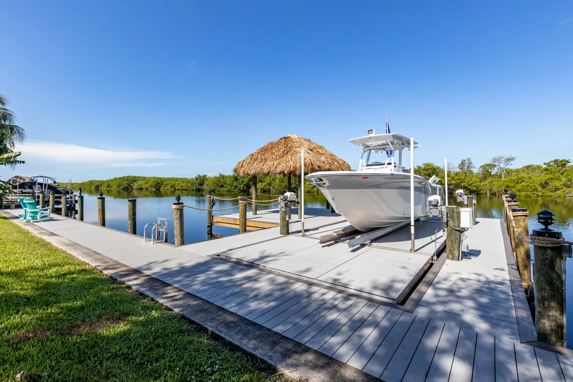 A dock with a tiki hut, boat on a lift, and mangroves in the background under a clear blue sky.