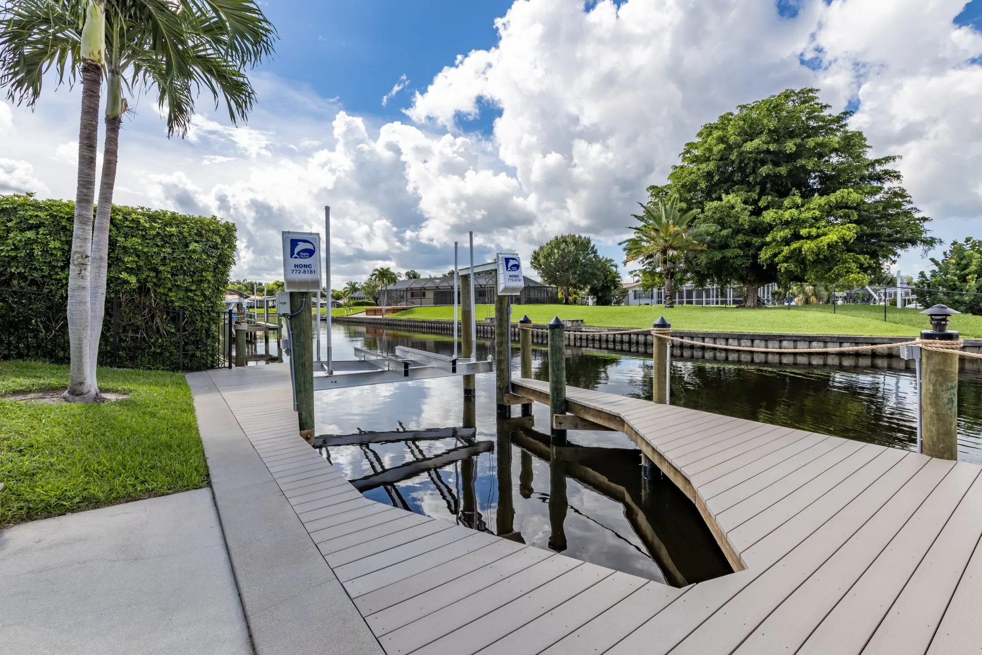 A curved dock walkway beside a canal with boat lifts, lush greenery, and clear skies in the background.