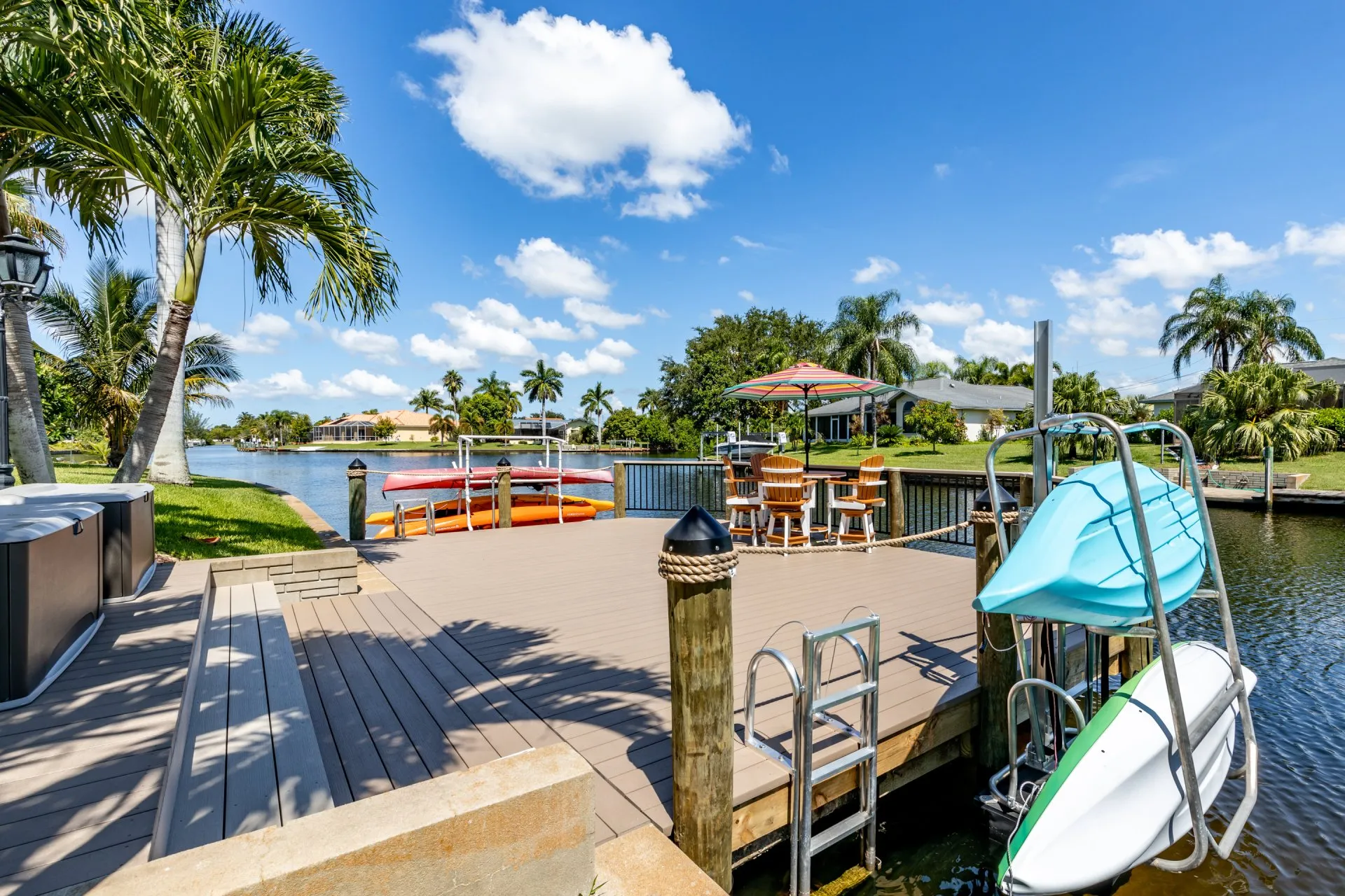 A dock featuring a kayak lift, ladder, Adirondack chairs, and a colorful umbrella on a sunny canal.