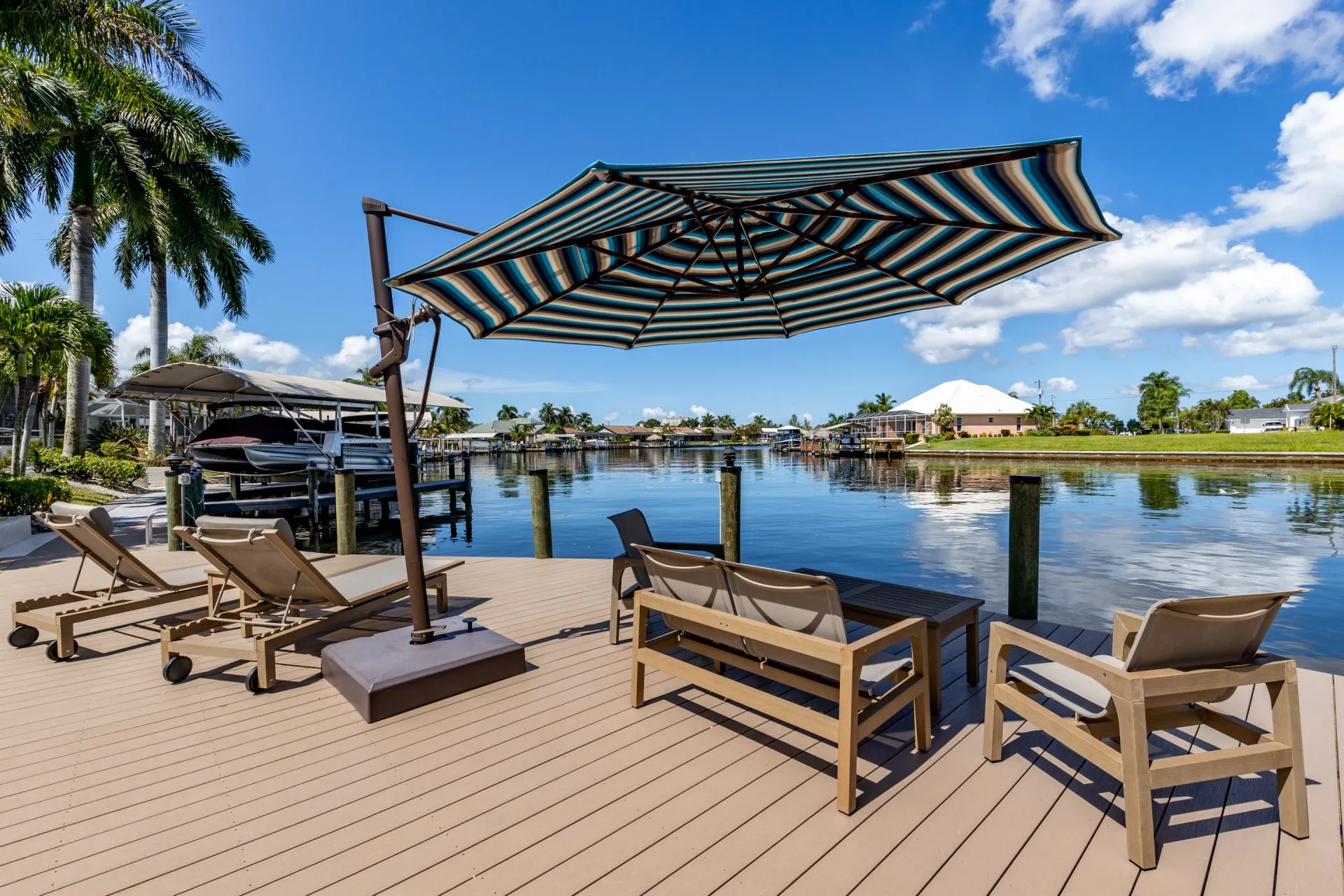 Waterfront dock with lounge chairs, a striped umbrella, and scenic views of palm trees and the canal.