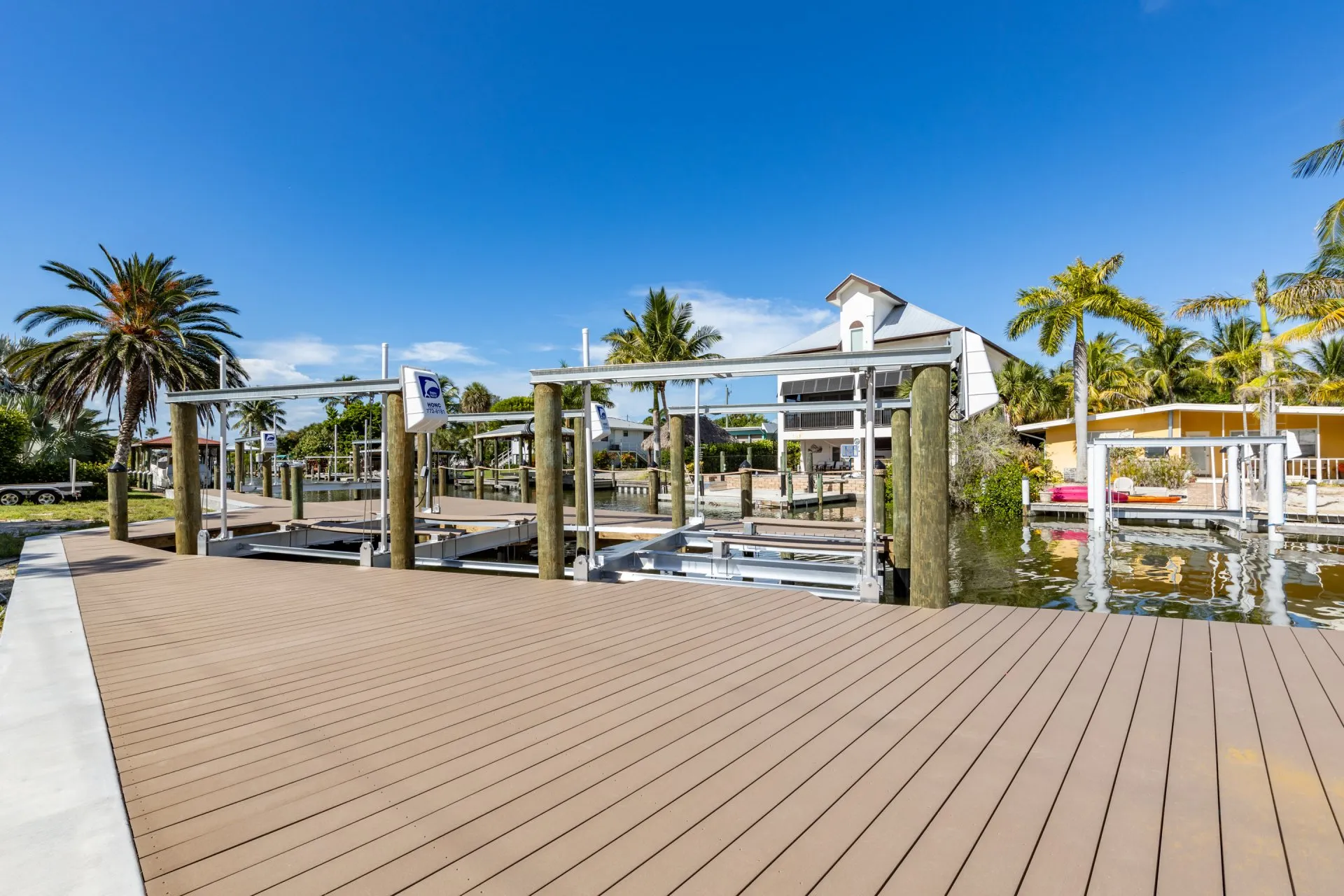 A waterfront dock featuring multiple boat lifts, surrounded by tropical homes and palm trees under a clear blue sky.