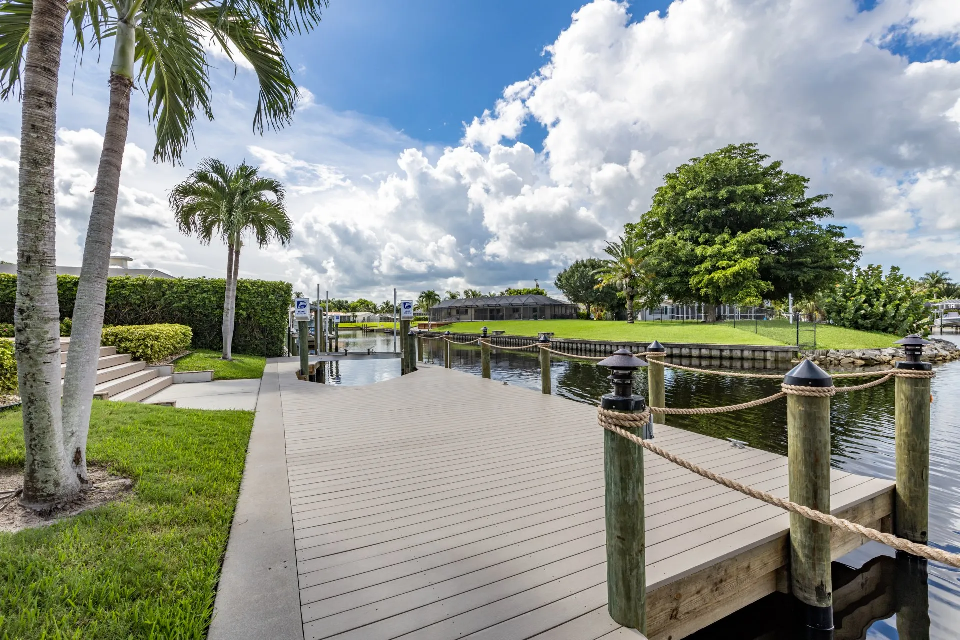 A dock with wooden posts and rope railing along a canal, surrounded by palm trees and greenery under a partly cloudy sky.