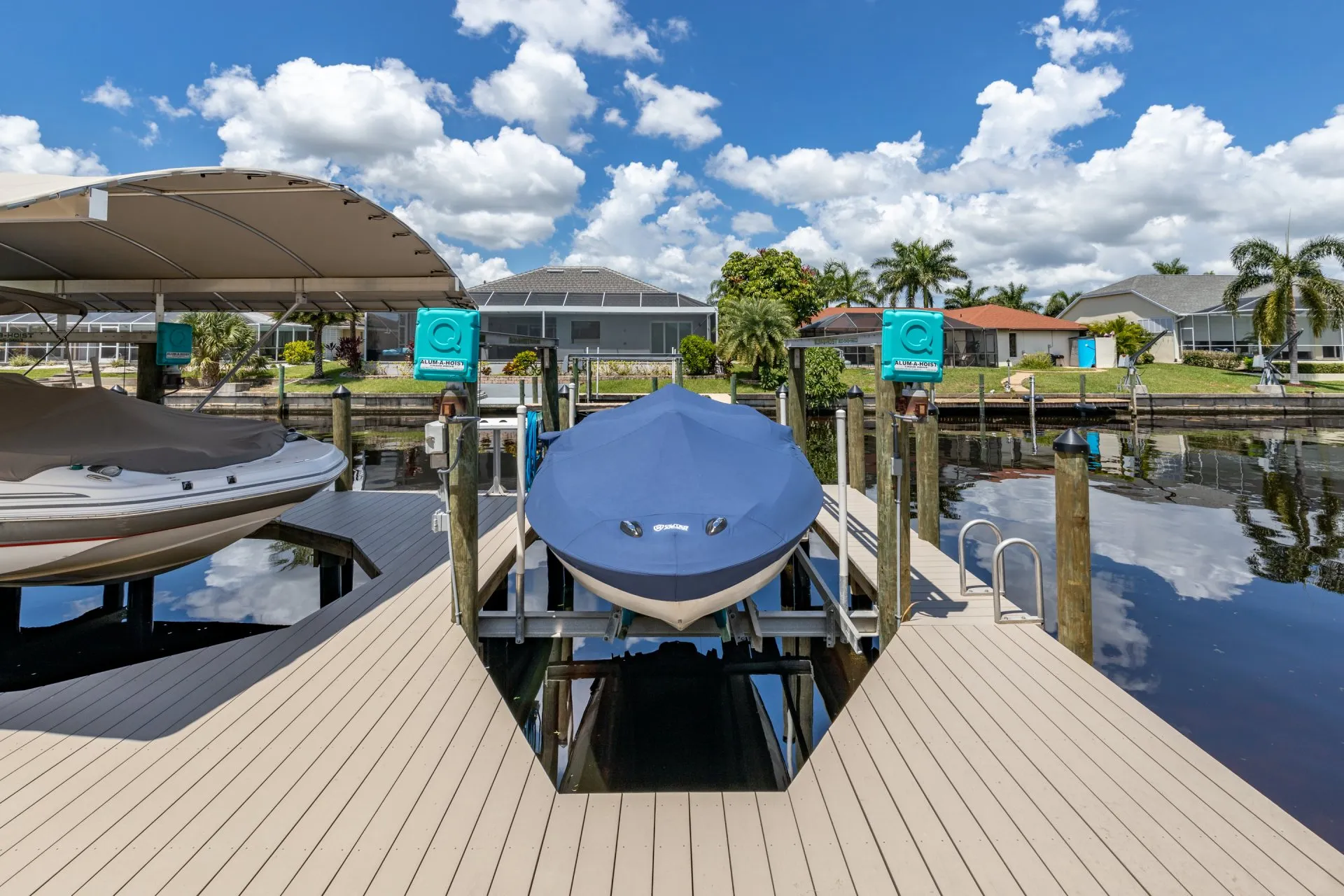 A covered boat on a lift at a dock, surrounded by a calm canal and residential homes.