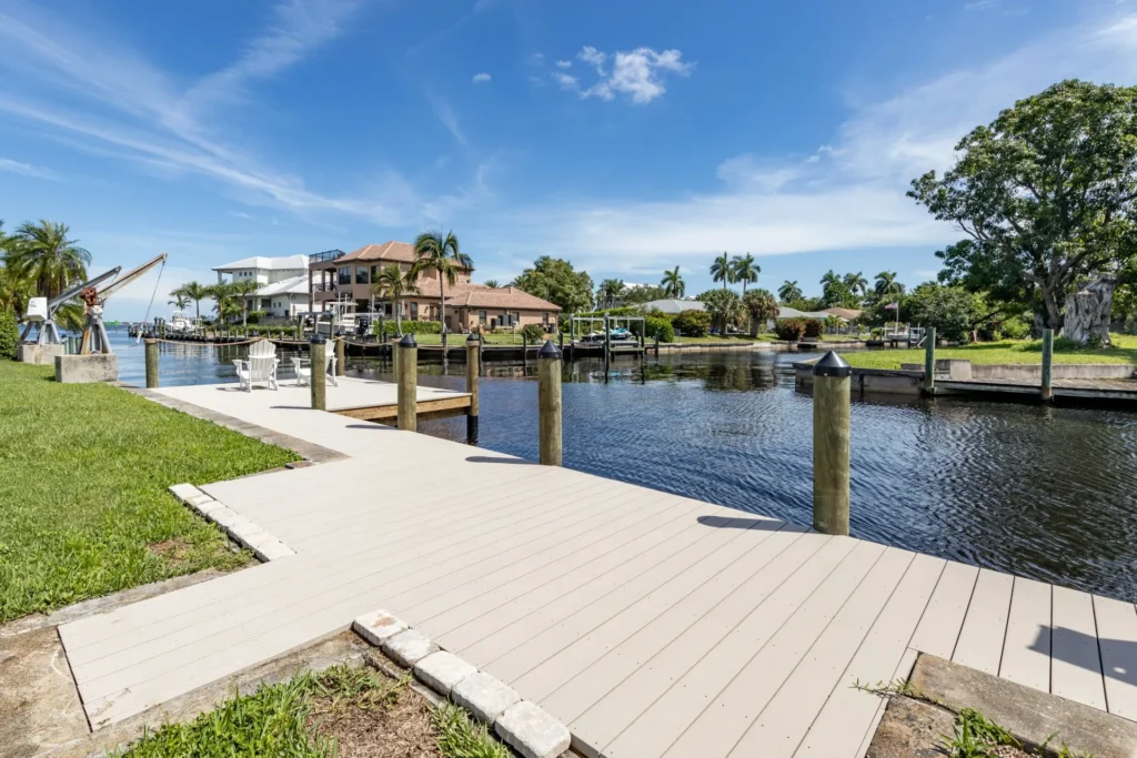 A wooden dock extending along a calm canal with houses and lush greenery in the background.