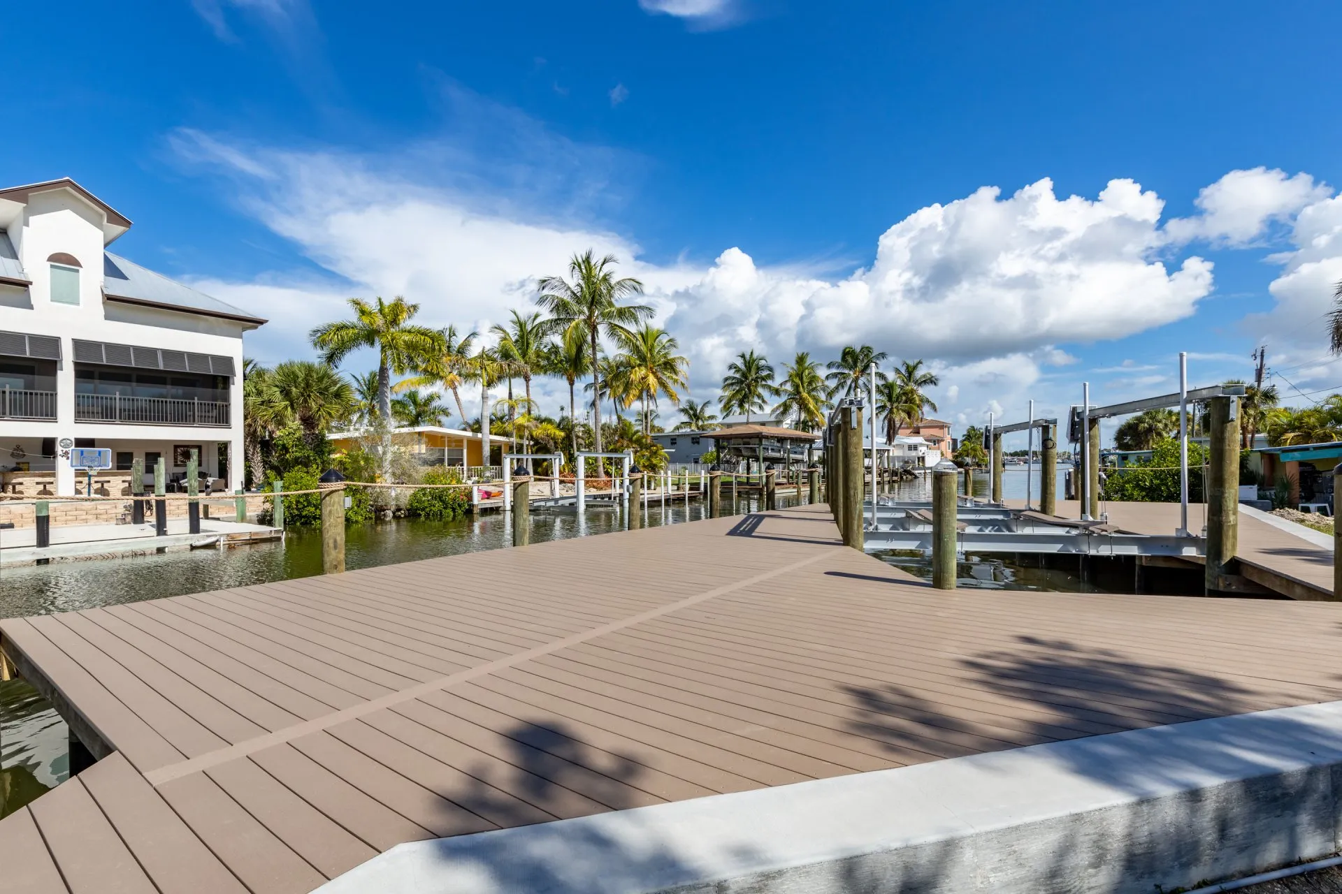 A large waterfront dock with boat lifts along a canal, surrounded by tropical homes and palm trees under a clear blue sky.