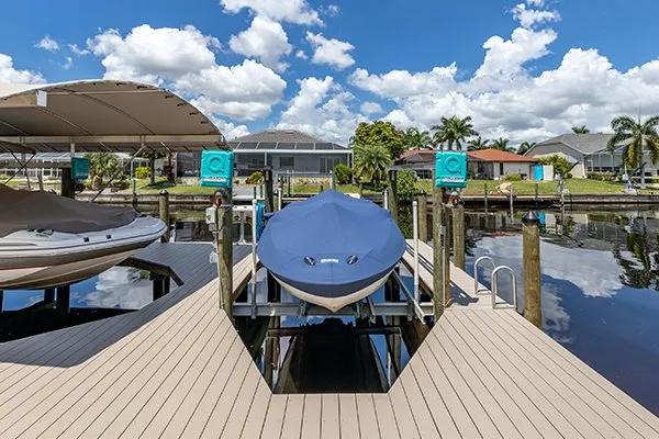 A front view of a boat on a covered lift, secured on a canal-side dock with a sunny residential backdrop.