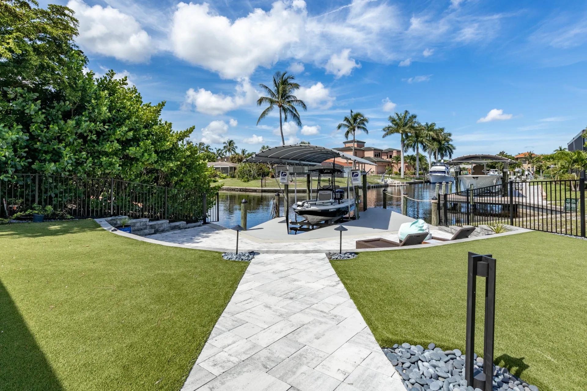 Landscaped backyard with a paved walkway leading to a boat lift on a canal surrounded by palm trees.