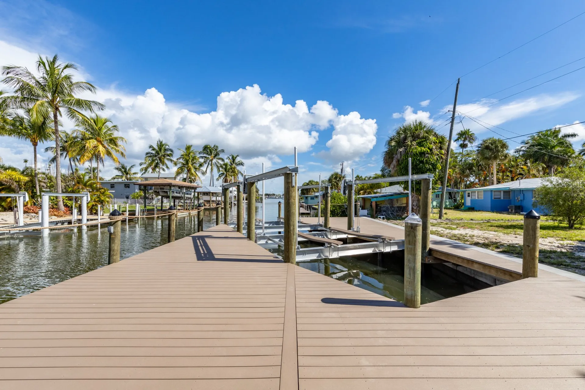 A spacious dock with boat lifts along a serene canal, surrounded by palm trees and tropical homes under a sunny sky.