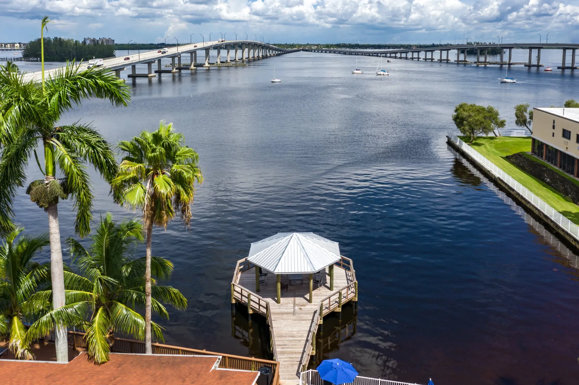 Aerial view of a riverside dock with a metal-roofed gazebo, surrounded by palm trees and overlooking a wide river with a bridge in the distance.