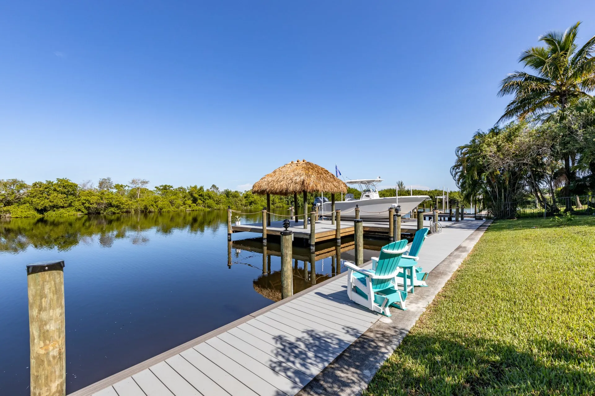 A dock with a tiki hut, boat lift, turquoise Adirondack chairs, and mangroves reflecting in calm water.