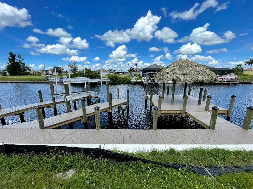 Wooden dock with a tiki hut and boat lift on calm waters, surrounded by lush greenery and blue skies.