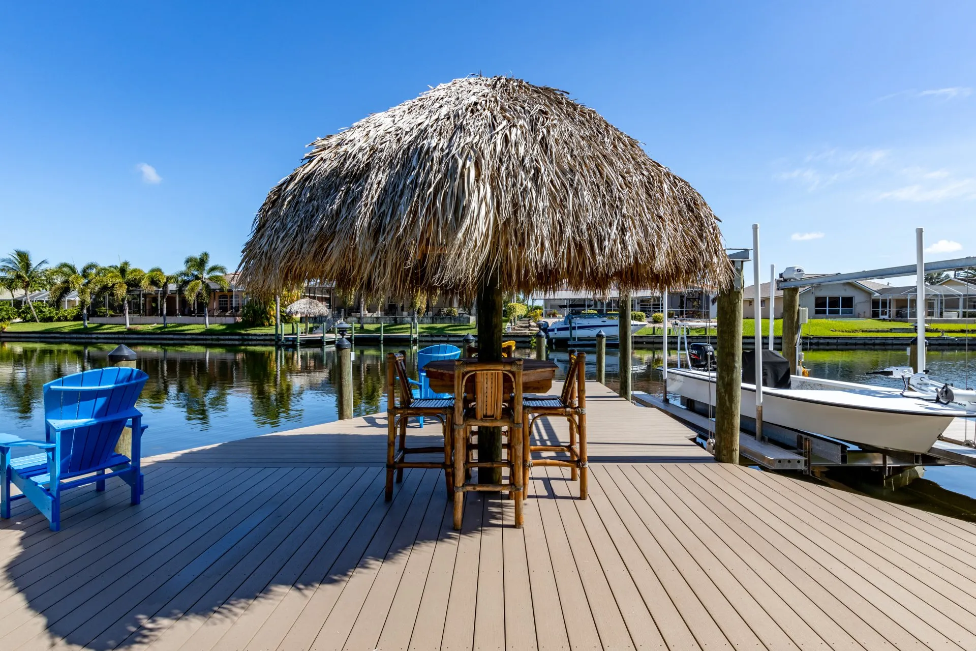 A waterfront dock featuring a tiki hut with bamboo furniture and blue Adirondack chairs under a bright blue sky.