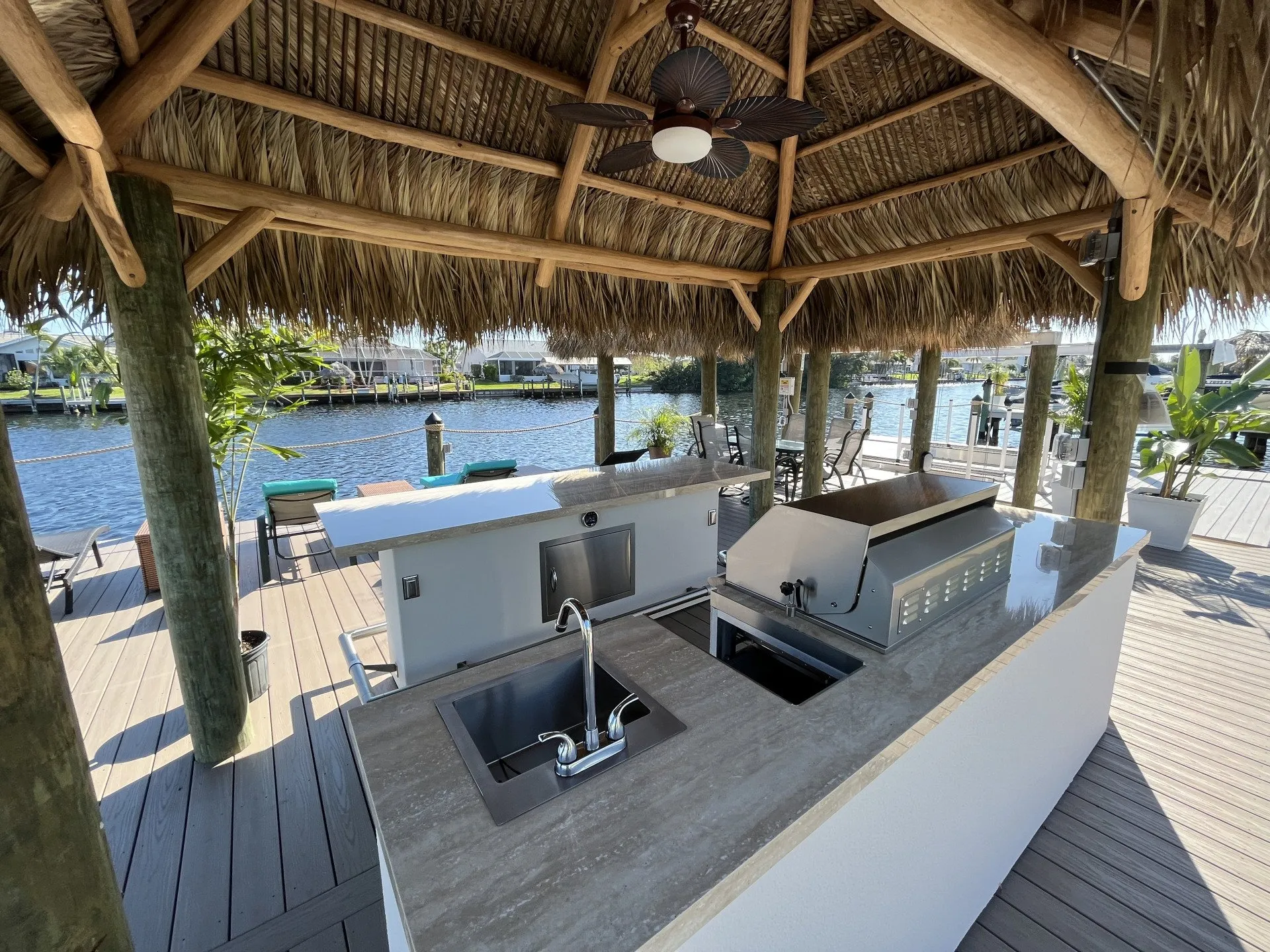 A modern outdoor kitchen under a tiki hut with a grill, sink, and spacious counter overlooking the water.