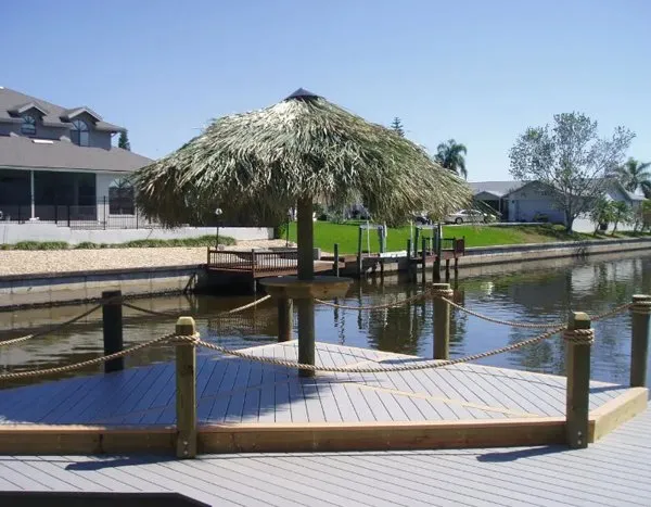 Tiki hut with a palm-thatch roof on a wooden dock, bordered by rope railings along a peaceful canal.