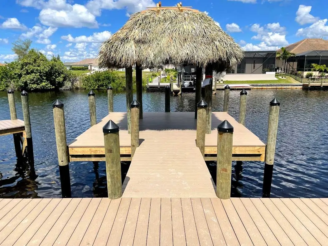 Wooden dock with a central tiki hut surrounded by water and wooden pilings under a bright blue sky.