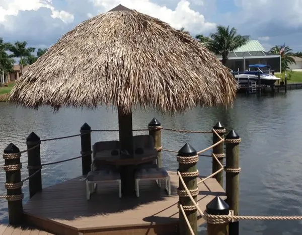 Tiki hut with a thatched roof on a wooden dock surrounded by rope railings along a calm canal.