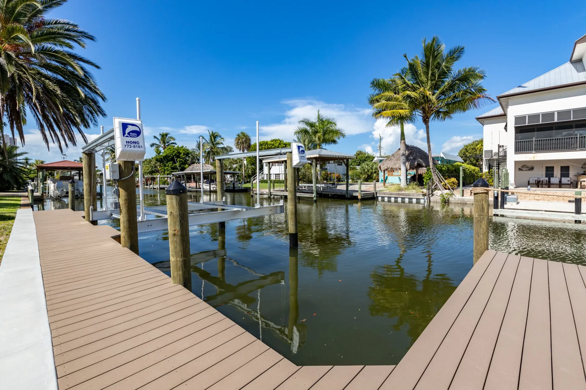 A waterfront dock with a boat lift, surrounded by palm trees and a tiki hut, in a serene canal setting.