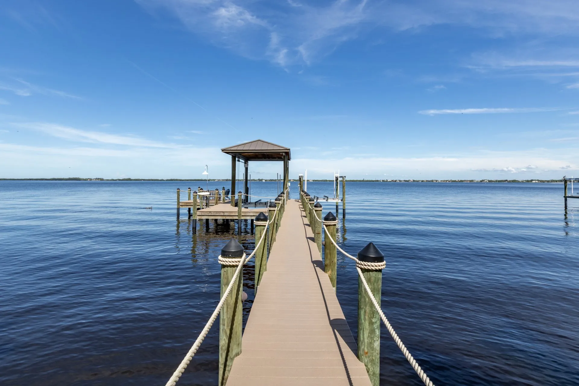 Long dock with rope railings extending over calm water to a covered seating area.