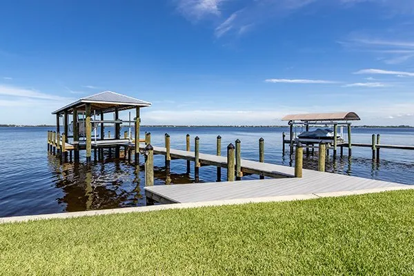 A waterfront dock with two covered boat lifts, calm waters, and a green lawn under a bright blue sky.