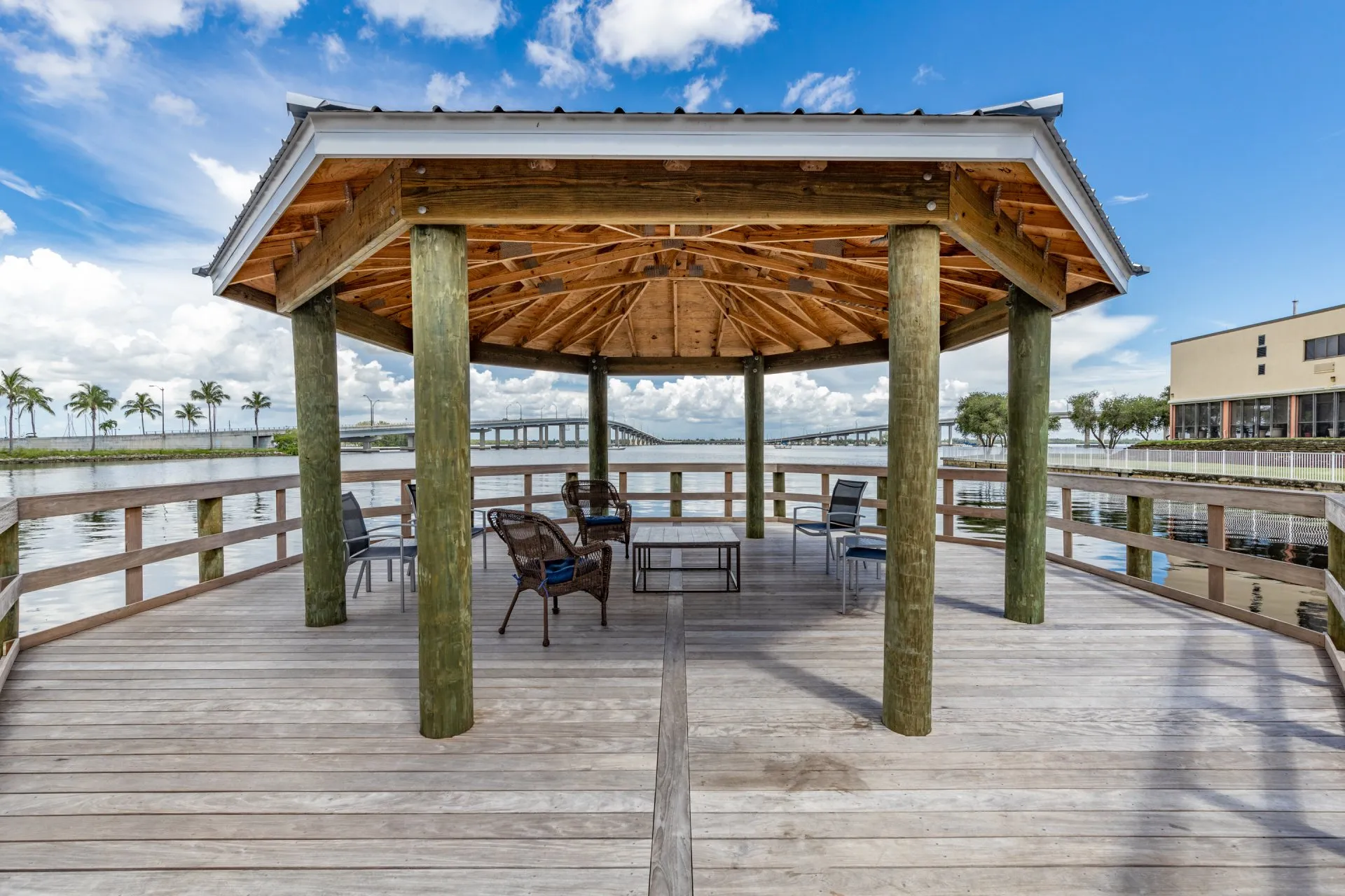 Waterfront gazebo with wooden beams and seating overlooking a canal and bridge.