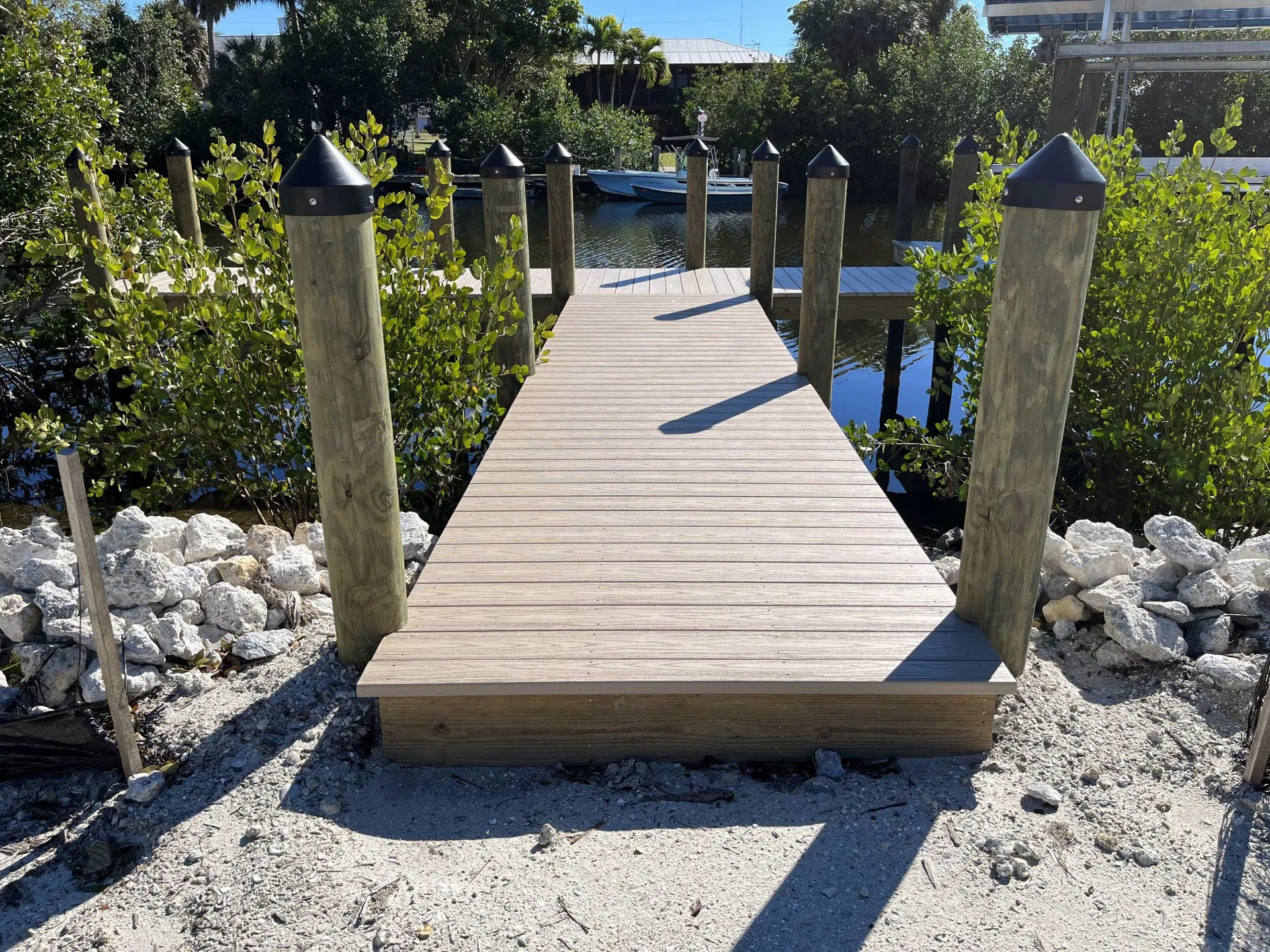 A wooden dock walkway with sturdy pilings leading to a calm canal, surrounded by mangroves and rocks.