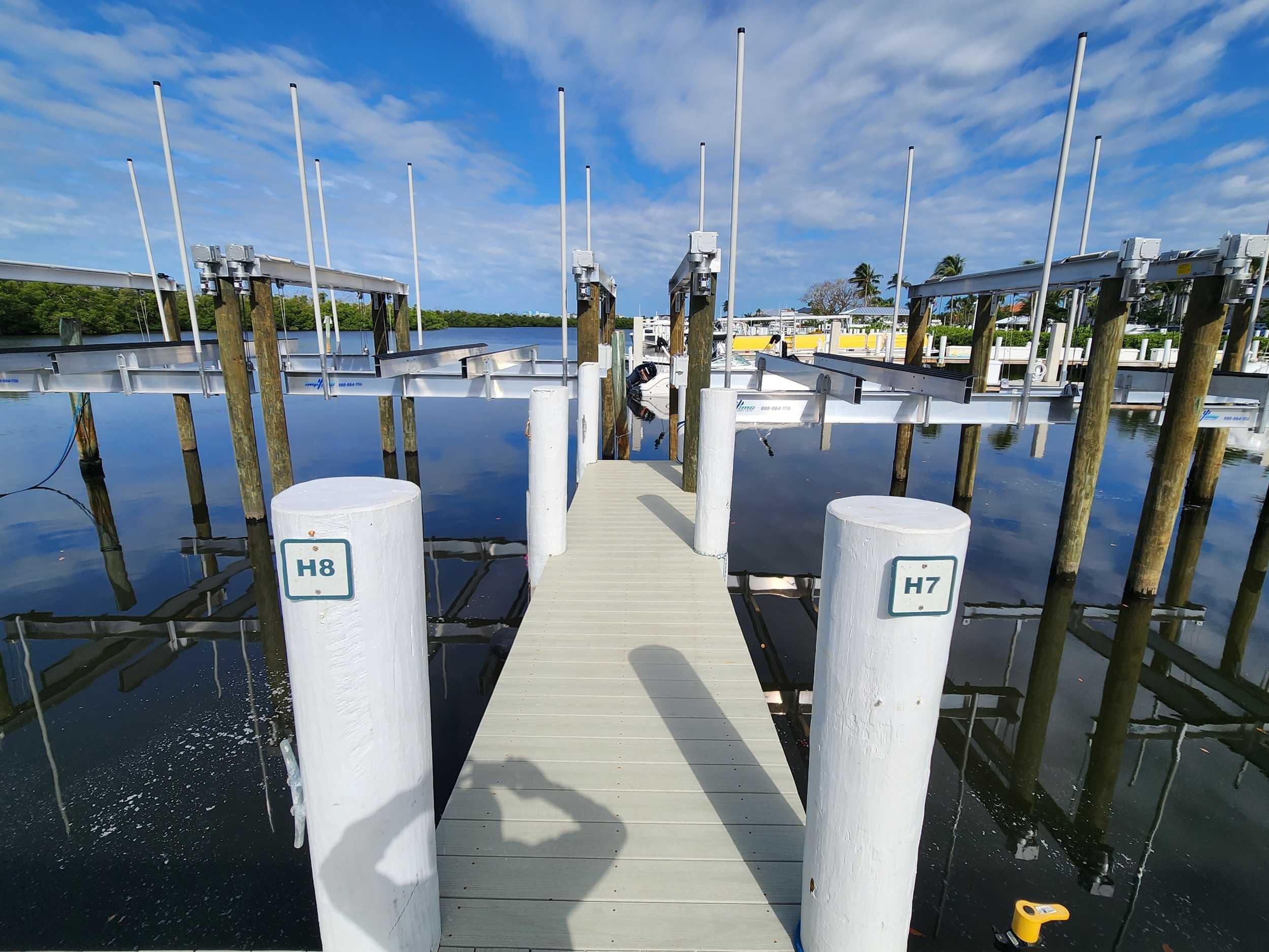 Boat slips with aluminum boat lifts at Port Sanibel Marina, viewed from a dock walkway between slips on calm water under a blue sky with scattered clouds.